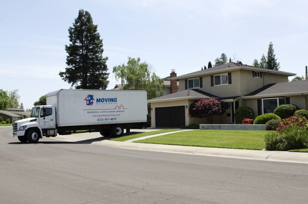 White moving truck with 'Moving With Grace' logo parked on a residential street in front of a beige two-story house with a green lawn and shrubs nearby.